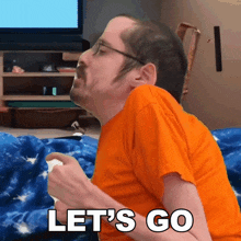 a man in an orange shirt says let 's go while holding a controller
