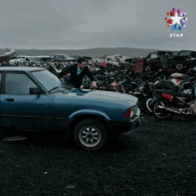 a blue car is parked in front of a pile of wrecked cars and motorcycles with a star in the background