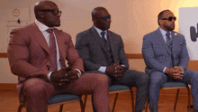 three men in suits sit in chairs with their arms crossed