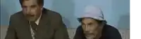 two men are sitting next to each other on a couch . one of the men is wearing a hat .