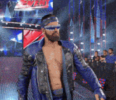 a shirtless wrestler in a blue jacket stands in front of a sign that says even