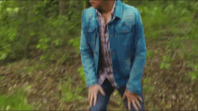 a man in a blue denim jacket and plaid shirt is walking through a forest .
