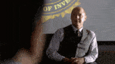 a man in a vest and tie sits in front of a sign that says ' department of investigation '