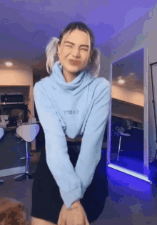 a woman wearing a blue sweater and a black skirt is standing in front of a mirror in a kitchen .