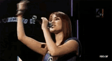 a woman singing into a microphone in front of a tv screen that says rbd.gif