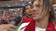 a man in a red shirt is talking into a microphone on a soccer field .