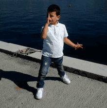 a young boy wearing a white polo shirt and jeans is standing in front of a body of water