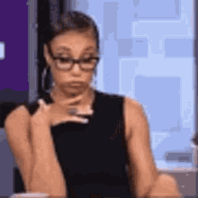 a woman wearing glasses and a black tank top is sitting at a table with her hands on her chin .