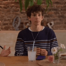 a young man in a striped sweater is sitting at a table with a glass of milk and a straw .