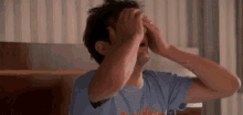 a man in a blue shirt is covering his face with his hands while sitting on a bed .