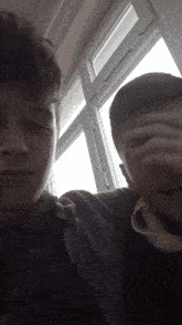 a man covering his face with his hand next to another man in front of a window