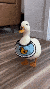 a white duck wearing a blue bib with a yellow duck on it is walking on the floor .