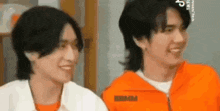 two young men are sitting next to each other and smiling . one of the men is wearing an orange jacket .