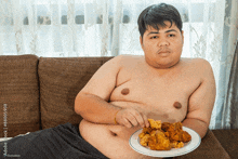 a shirtless man sits on a couch with a plate of fried chicken