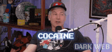 a man in a hat is sitting in front of a microphone with the words cocaine dark web behind him .