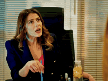a woman in a blue jacket sits at a desk with a bottle of lemonade in front of her