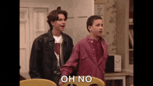 two young men are standing next to each other in a living room . one of the men is wearing a leather jacket .