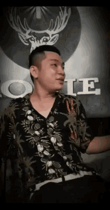 a man in a hawaiian shirt is sitting in front of a sign with a deer head on it .