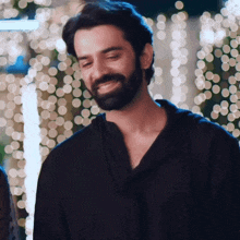 a man with a beard wearing a black shirt smiles in front of christmas lights