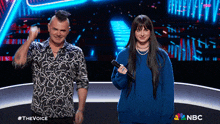 a man with a mohawk and a woman in a blue sweater stand on a stage in front of a nbc sign