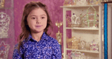 a little girl wearing a blue shirt is standing in front of a shelf filled with tiaras .