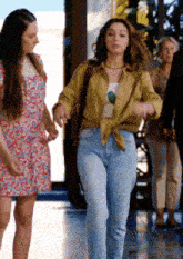 a woman in a yellow shirt and blue jeans is walking with another woman in a floral dress .