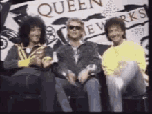 three men are sitting next to each other in front of a sign that says queen new works .