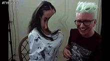 a man with green hair and glasses is sitting next to a woman with a large breast .