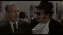 two men in suits and hats are talking to each other . one of the men is wearing sunglasses .