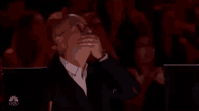 a man in a suit and glasses is covering his mouth with his hand while sitting in a theatre .