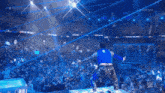 a man in a blue shirt is standing on a stage in front of a crowd with peacock in the background