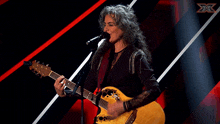 a woman singing into a microphone while holding a guitar in front of an x logo