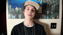 a young man wearing a hat and a necklace stands in front of a cityscape painting