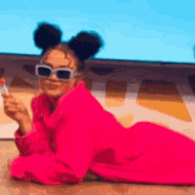 a woman wearing a pink robe and sunglasses is laying on the floor holding a lollipop .