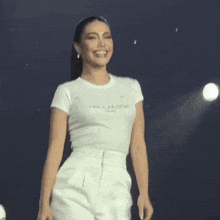 a woman wearing a white t-shirt and white pants is smiling on a stage