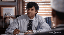 a man in a striped shirt and tie sits at a desk with a pen in his hand and says snap
