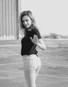 a woman in a black shirt and white jeans is standing in a parking lot in a black and white photo .