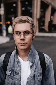 a young man wearing glasses and a denim jacket is standing on a street