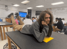 a man laying on a desk in a classroom with a poster on the wall that says periodic table of the elements