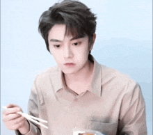 a young man in a tan shirt is eating with chopsticks .