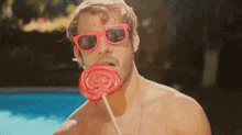 a shirtless man wearing sunglasses is eating a large lollipop .