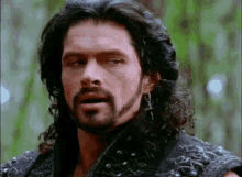 a man with long hair and a beard is standing in the woods and making a funny face .