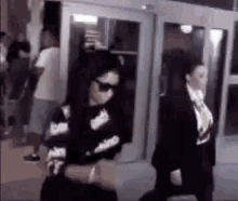 a woman wearing sunglasses is walking out of a building with a man in a suit behind her .