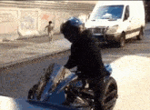 a man riding a motorcycle on a street with a white van in the background and a wall with graffiti on it that says cars