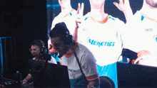 a man wearing a heretics shirt stands in front of a computer screen