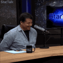 a man sitting at a table with a microphone in front of a screen that says startalk