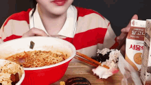 a woman in a red and white striped shirt is eating a bowl of noodles and drinking milk .