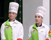 two women wearing chef hats and jackets are standing next to each other in a room .