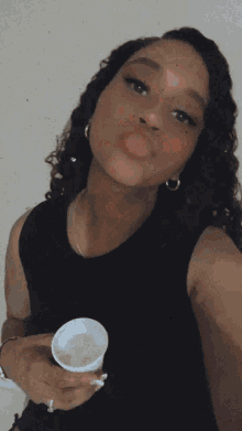 a woman in a black tank top is holding a white container and sticking her tongue out