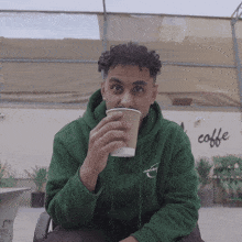 a man in a green hoodie drinking from a cup with the word coffee on the wall behind him